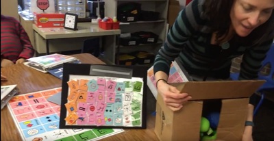 Educator working with learners using a box with engaging materials to create opportunities for communication. AAC systems are visible in the environment, and the educator is only giving a peek at the fun things in the box. She will model AAC to support learners to participate authentically.